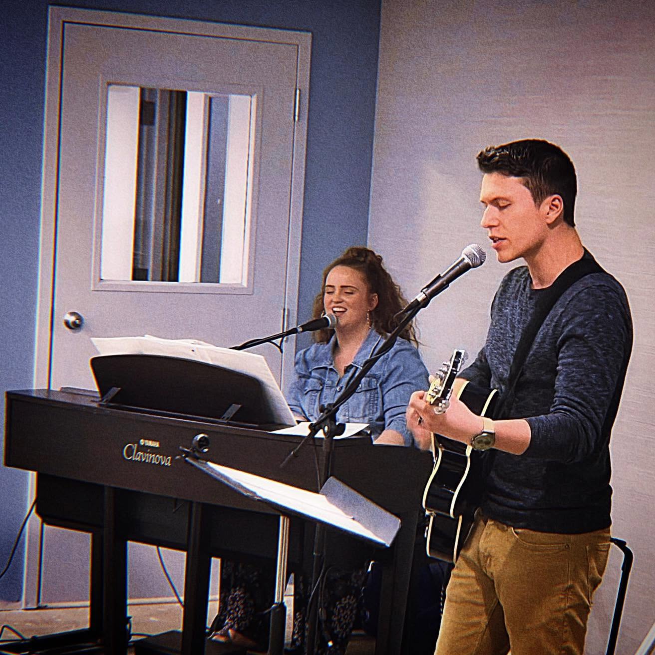 A man plays guitar and sings into a microphone, while a woman sings into a microphone and plays keyboard in a room.