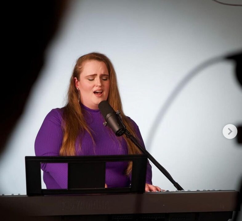 Woman singing into a microphone at a keyboard, wearing a purple top, with a plain background.
