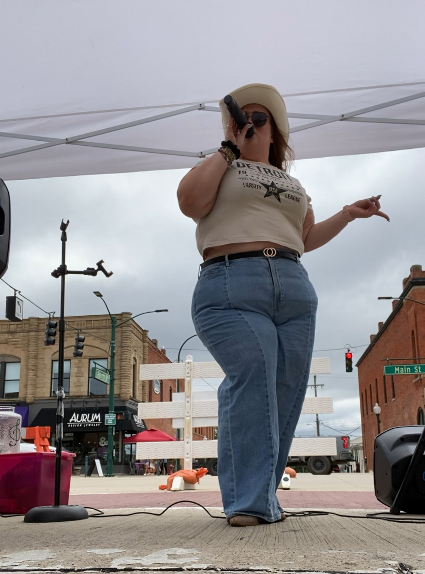 Woman standing outdoors under a canopy, wearing a hat, sunglasses, a beige shirt, and blue jeans, with buildings and street in background.