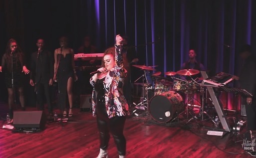 Performer singing on stage with backup singers and musicians, dark curtain backdrop, wooden floor, stage lighting.
