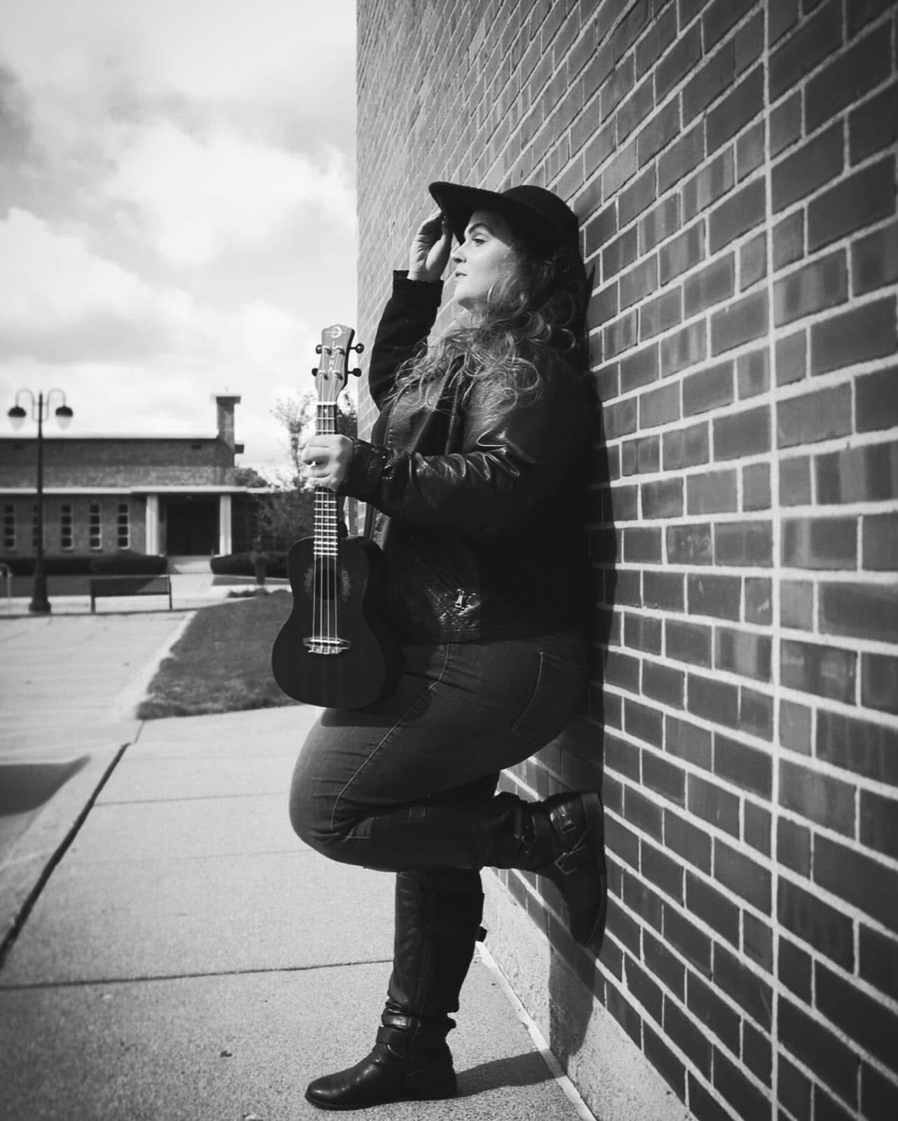 Person leaning against a brick wall holding a ukulele, wearing a hat and jacket, outdoors on a sidewalk.