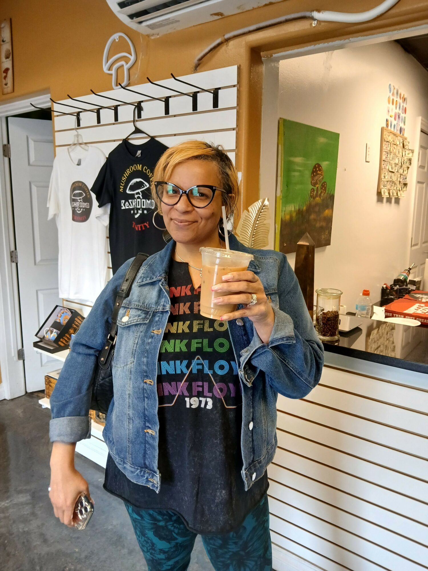 Person with glasses and blonde hair holding a phone, wearing a denim jacket and black T-shirt, standing indoors.