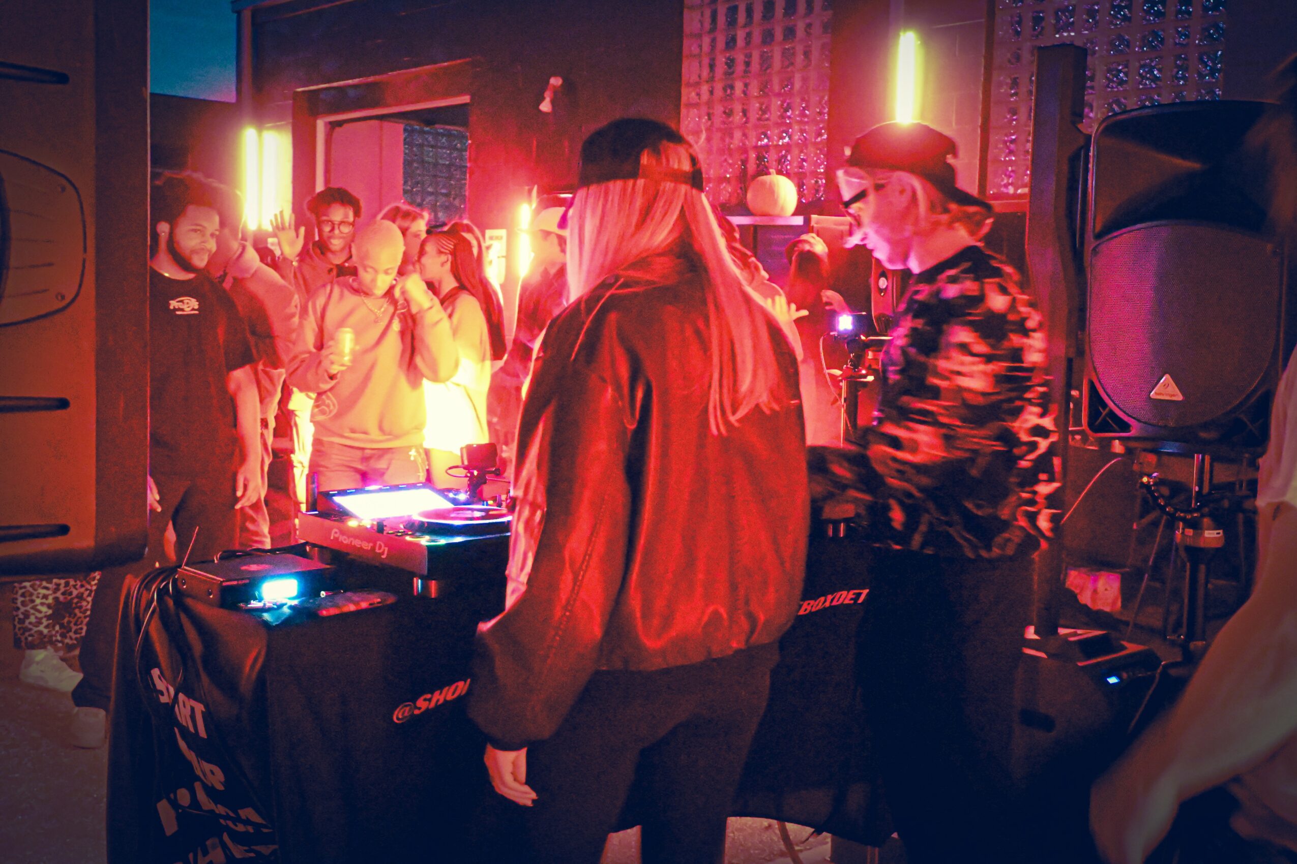 People dancing and socializing in a nightclub with colorful lighting and a DJ setup, lively atmosphere, and a person in a hat.