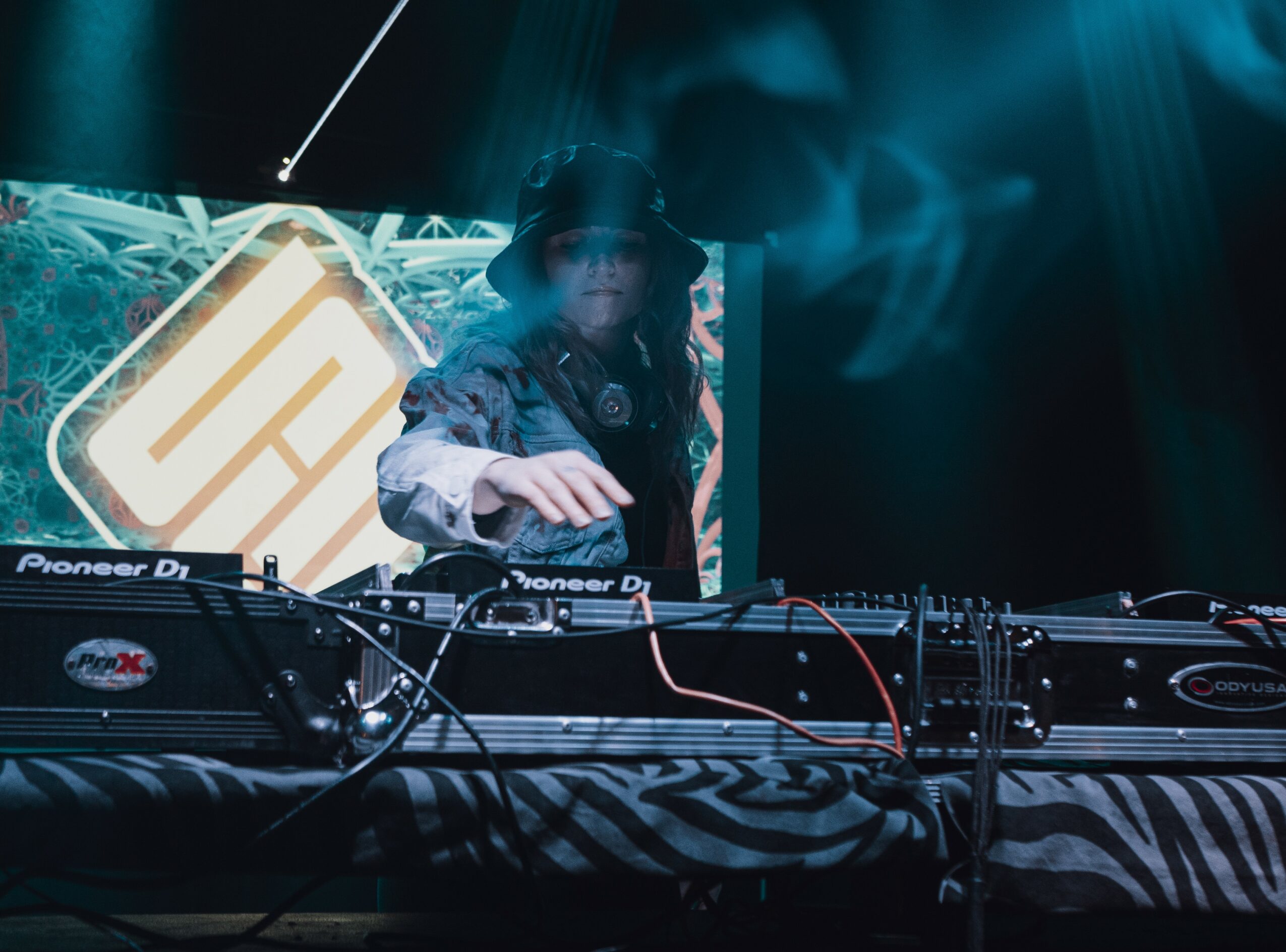DJ wearing a hat and headphones, operating equipment on stage with a digital display behind.