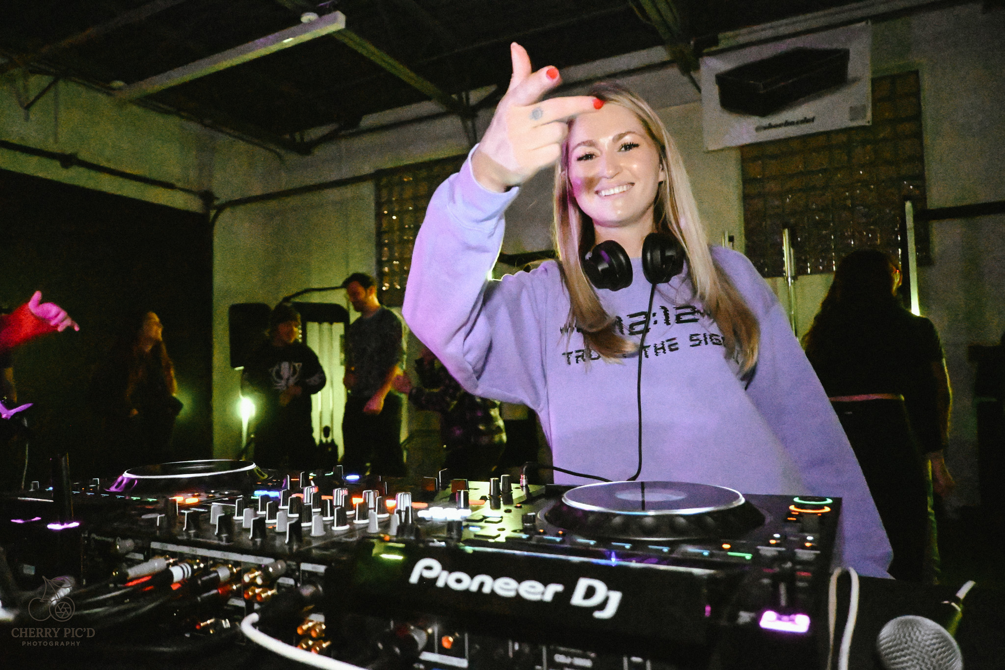 Female DJ smiling and giving a thumbs-up at a music event with DJ equipment and people in the background.