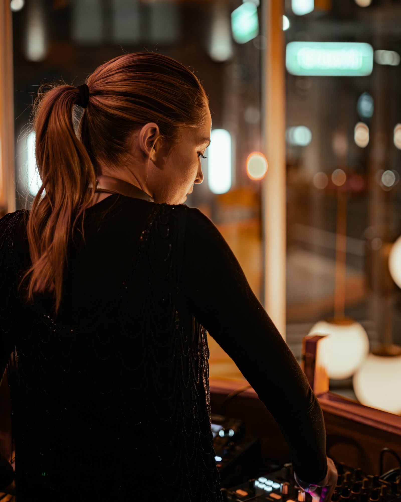 Woman with a ponytail looking down at a control panel in a dimly lit indoor setting, city lights visible outside.