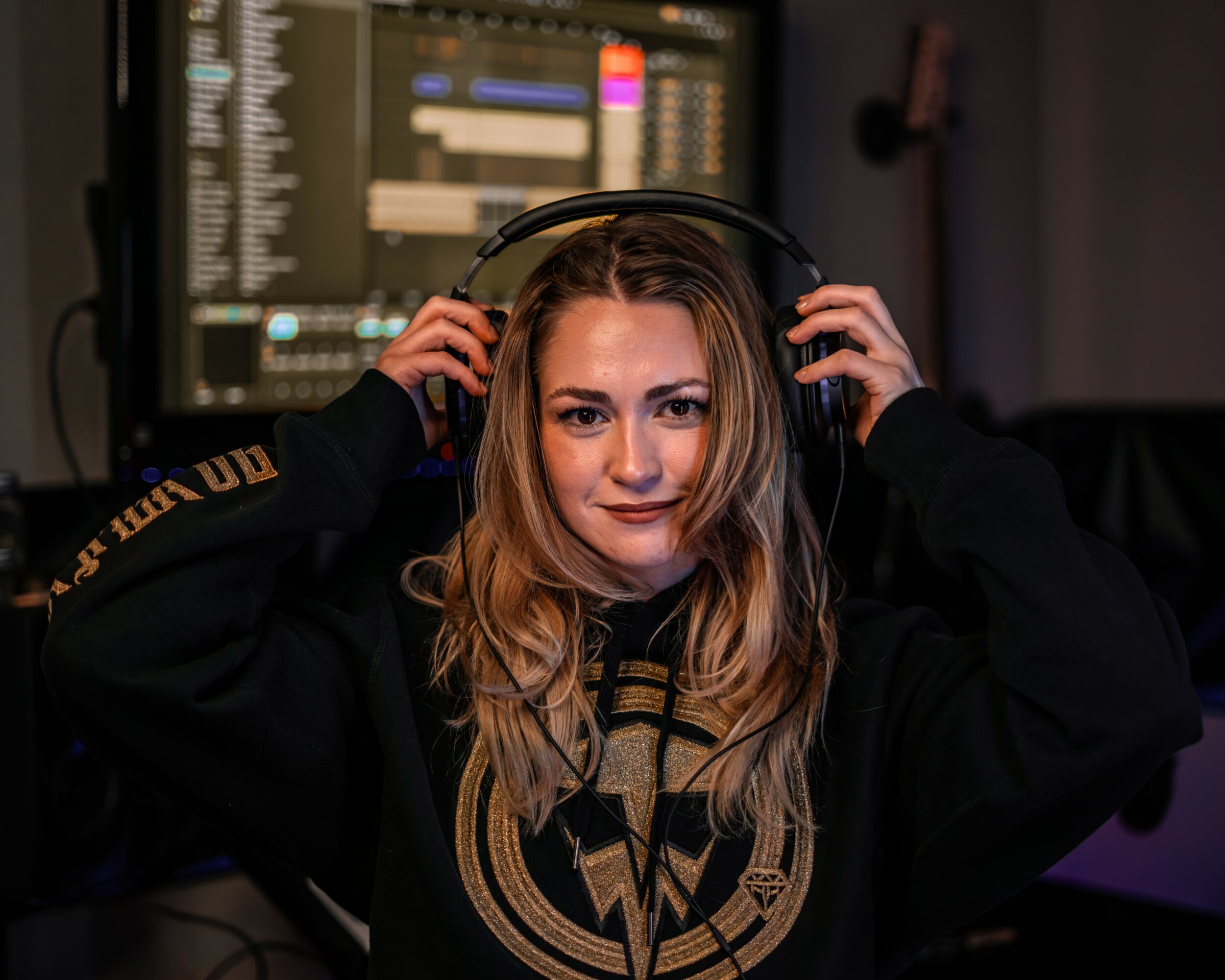 Young woman with headphones in a music studio, computer screen in background, smiling, wearing a black hoodie with a gold emblem.