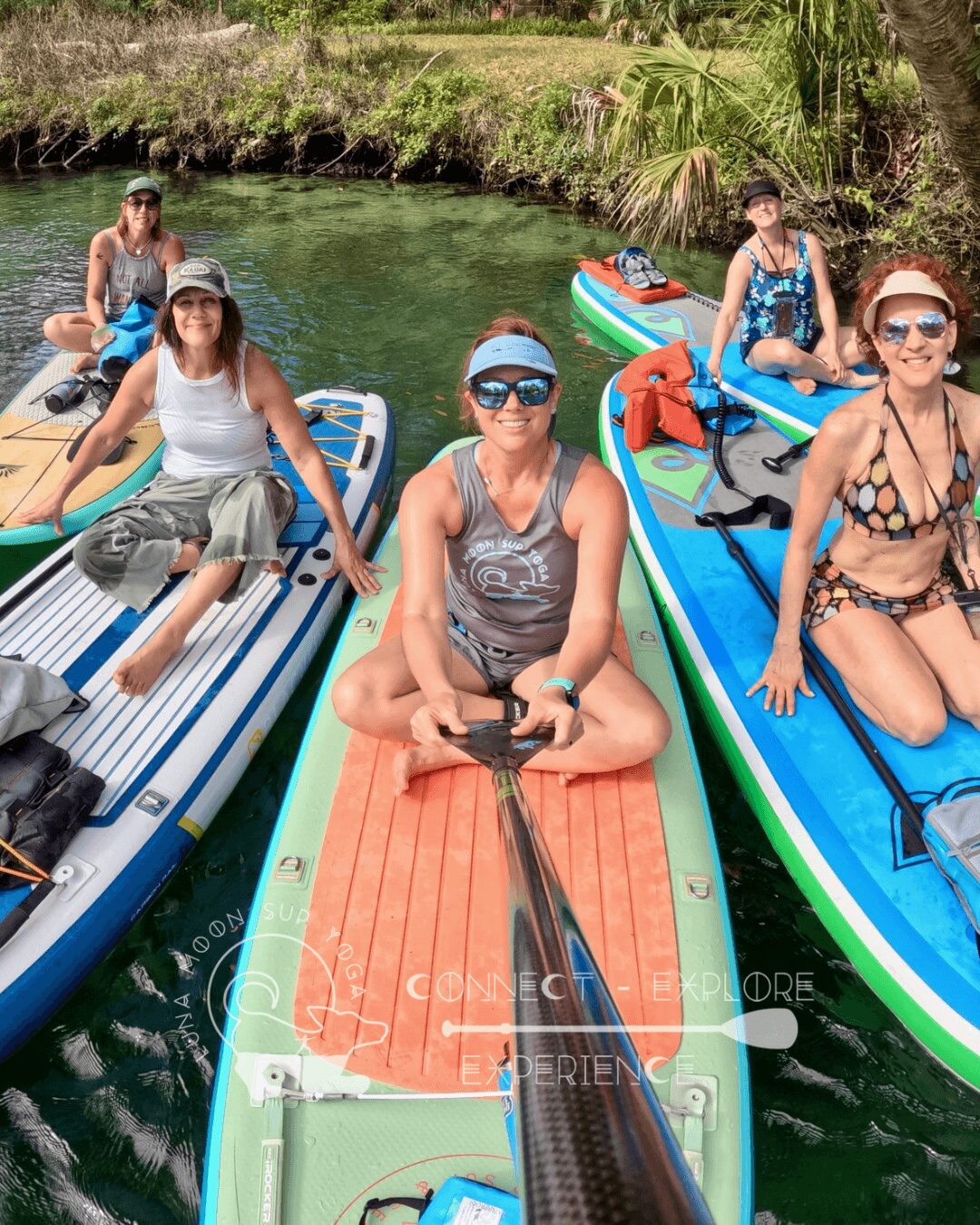 Group of six people on paddleboards in a river, some sitting and some standing, with greenery in background.