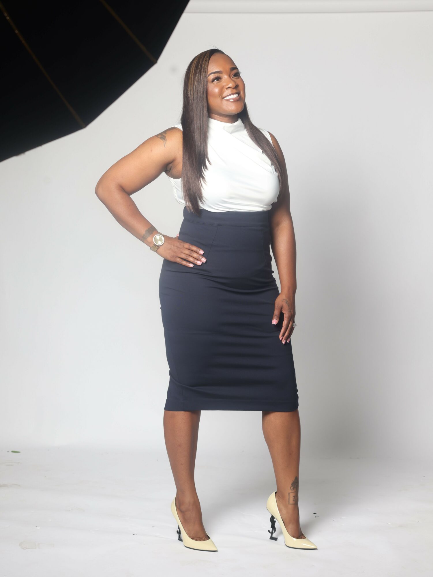 Woman in a black dress and white top standing against a plain background, smiling, with her right hand on her hip.