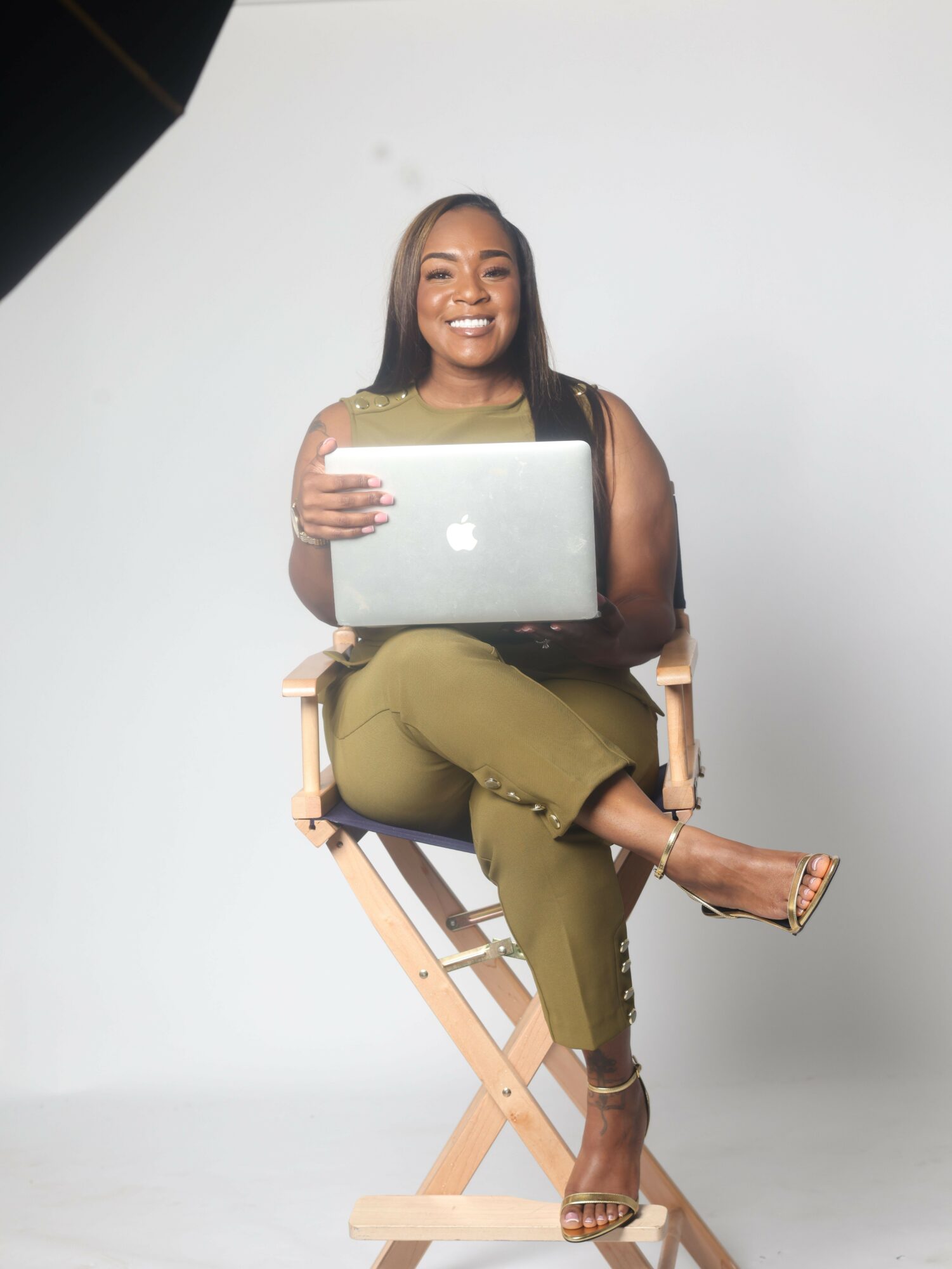 Woman sitting on a wooden chair holding a laptop, smiling, against a plain white background.