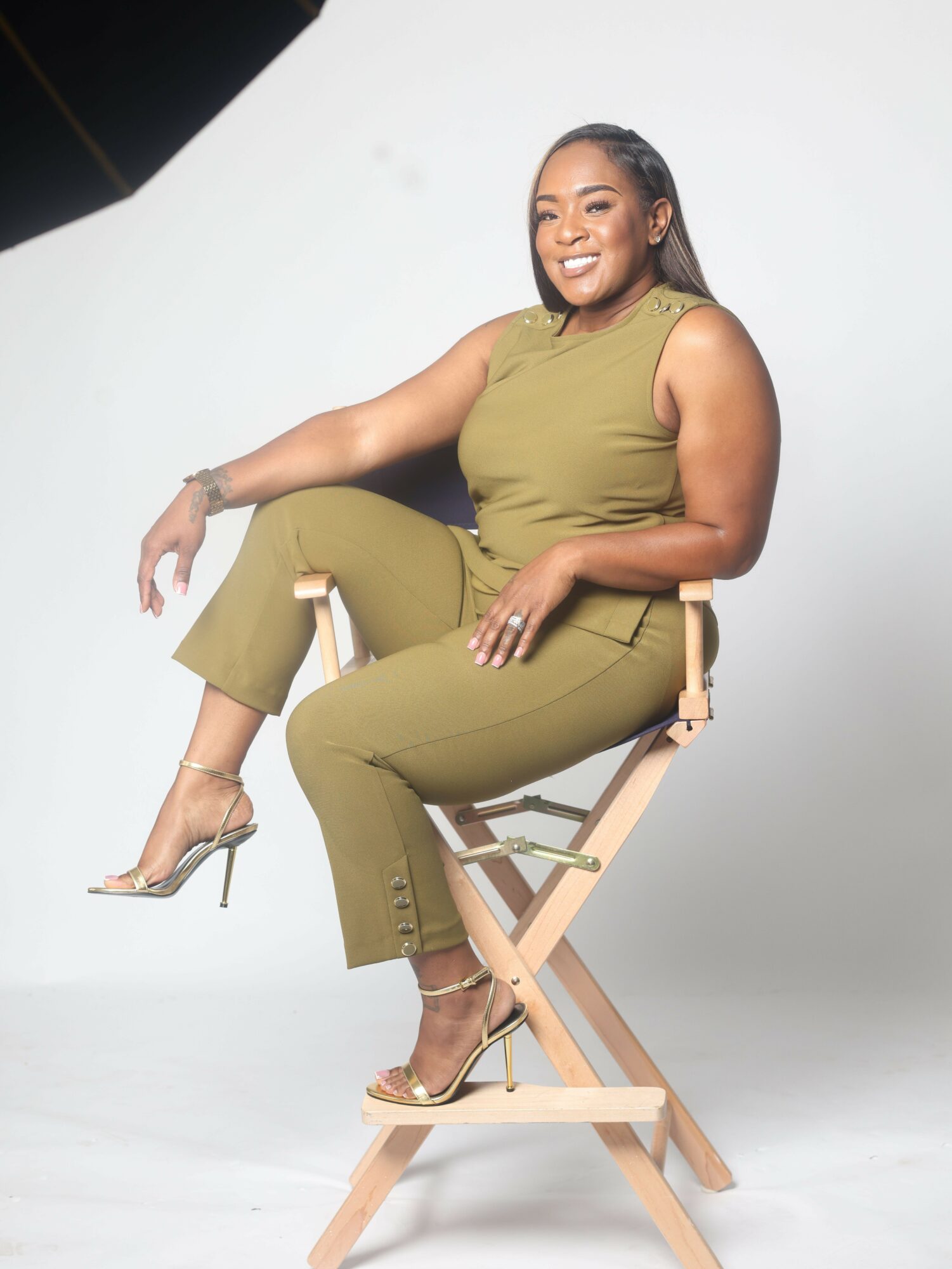 Woman sitting on a wooden chair, smiling, wearing a green outfit and high heels, against a plain white background.