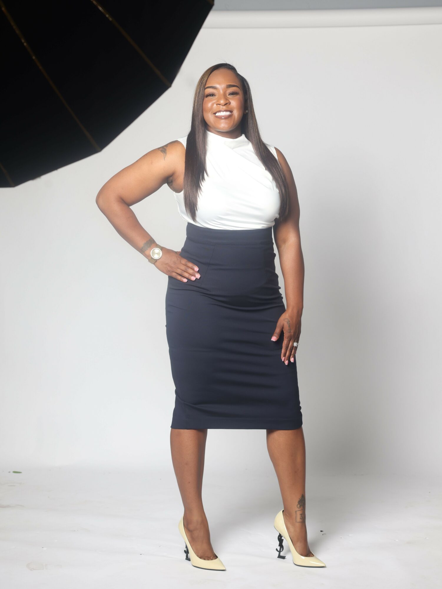 Woman in a black dress and white top standing against a plain background, smiling, with long hair and beige shoes.