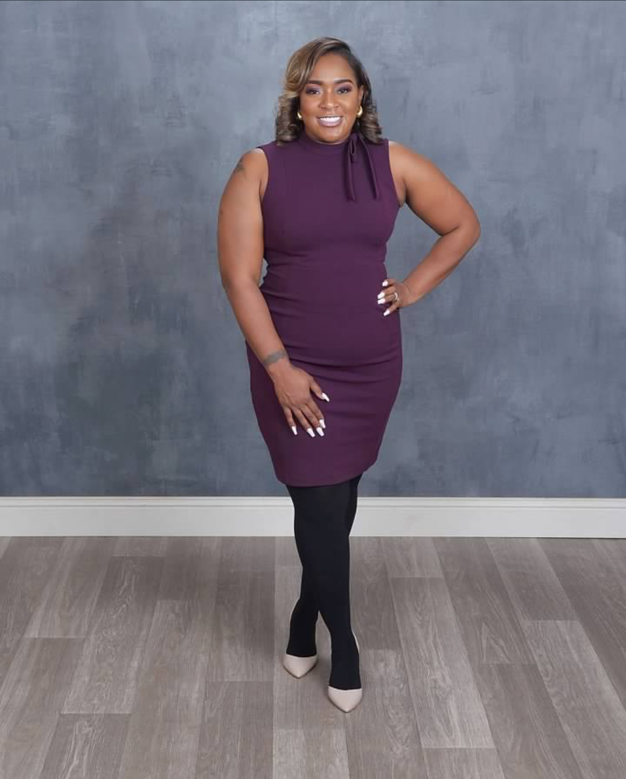 Woman in purple dress and black tights standing with hand on hip, smiling, against gray wall background.