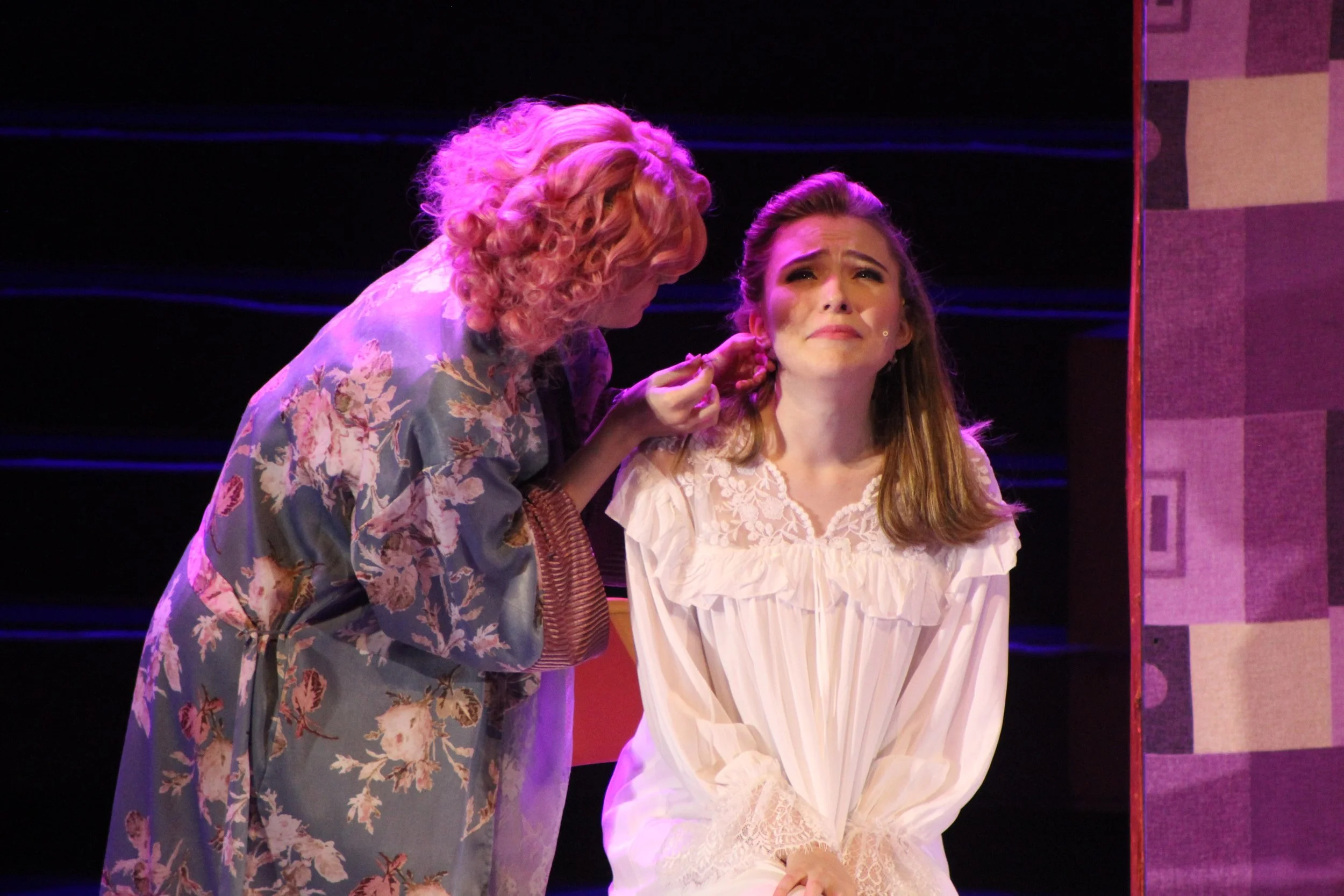 Two women on stage, one with curly hair leaning close to the other with straight hair, who appears upset.