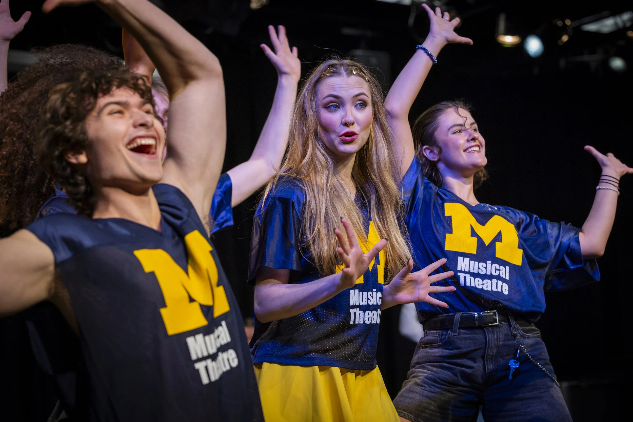Three young women on stage, wearing blue shirts with a yellow 'M' and 'Music Theatre' text, performing with expressive gestures.