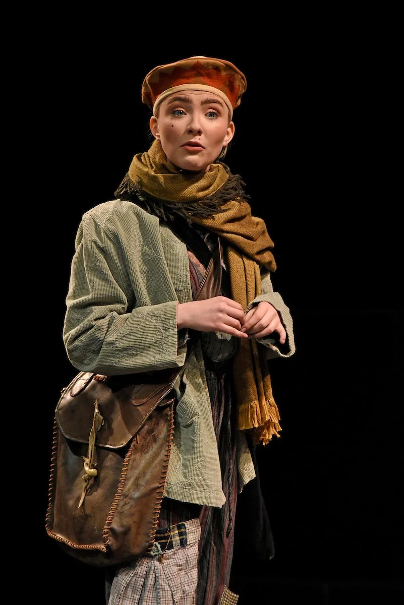 Woman with red hair wearing a green jacket and scarf, holding her hands together, against a black background.
