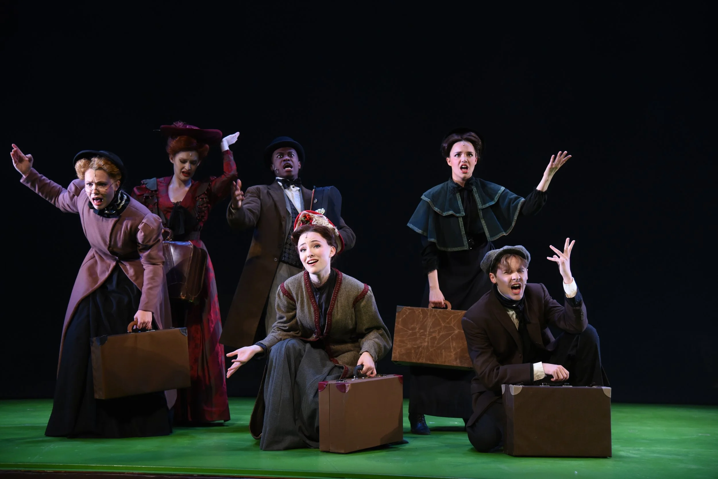 Six people on stage with boxes, performing a theatrical scene, dark background, green floor, expressive gestures.