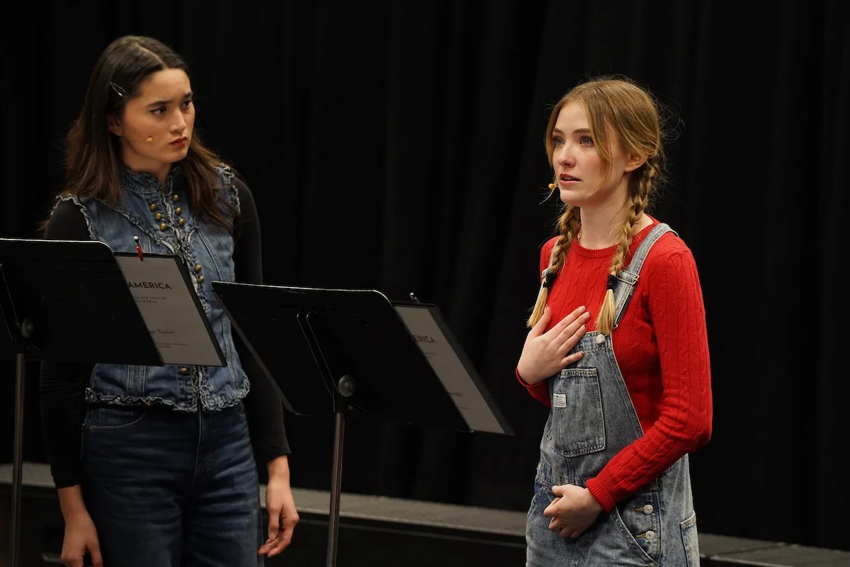 Two women standing and talking in front of black curtains, with music stands in front of them.