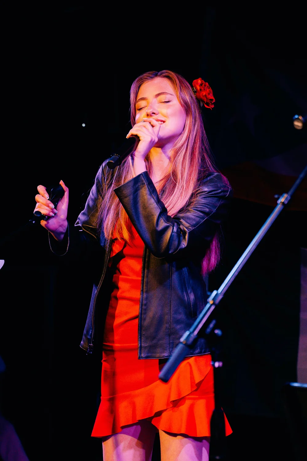 Young woman singing into a microphone on stage, wearing a red dress and black jacket, with a red flower in her hair.