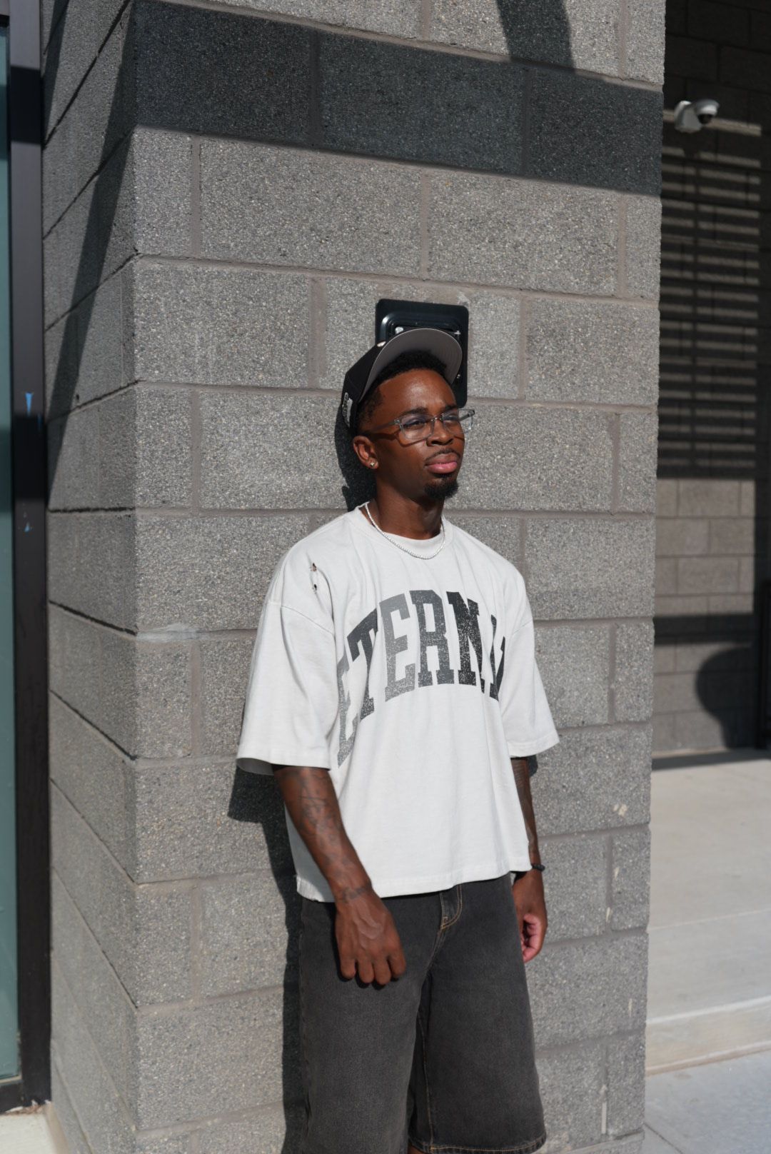 Person standing against a gray stone wall, wearing sunglasses, a white T-shirt, and dark pants, holding a phone.