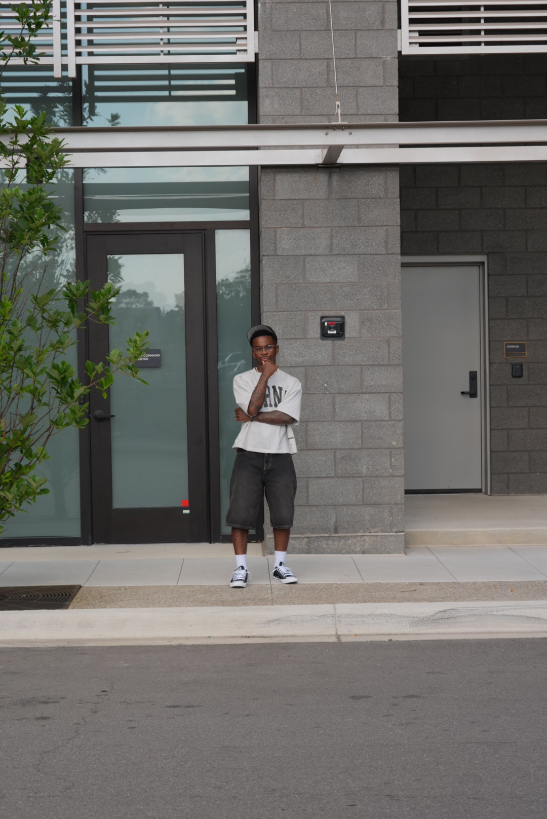 Young person standing outside a modern building with glass door and gray brick wall, holding a phone to their ear.