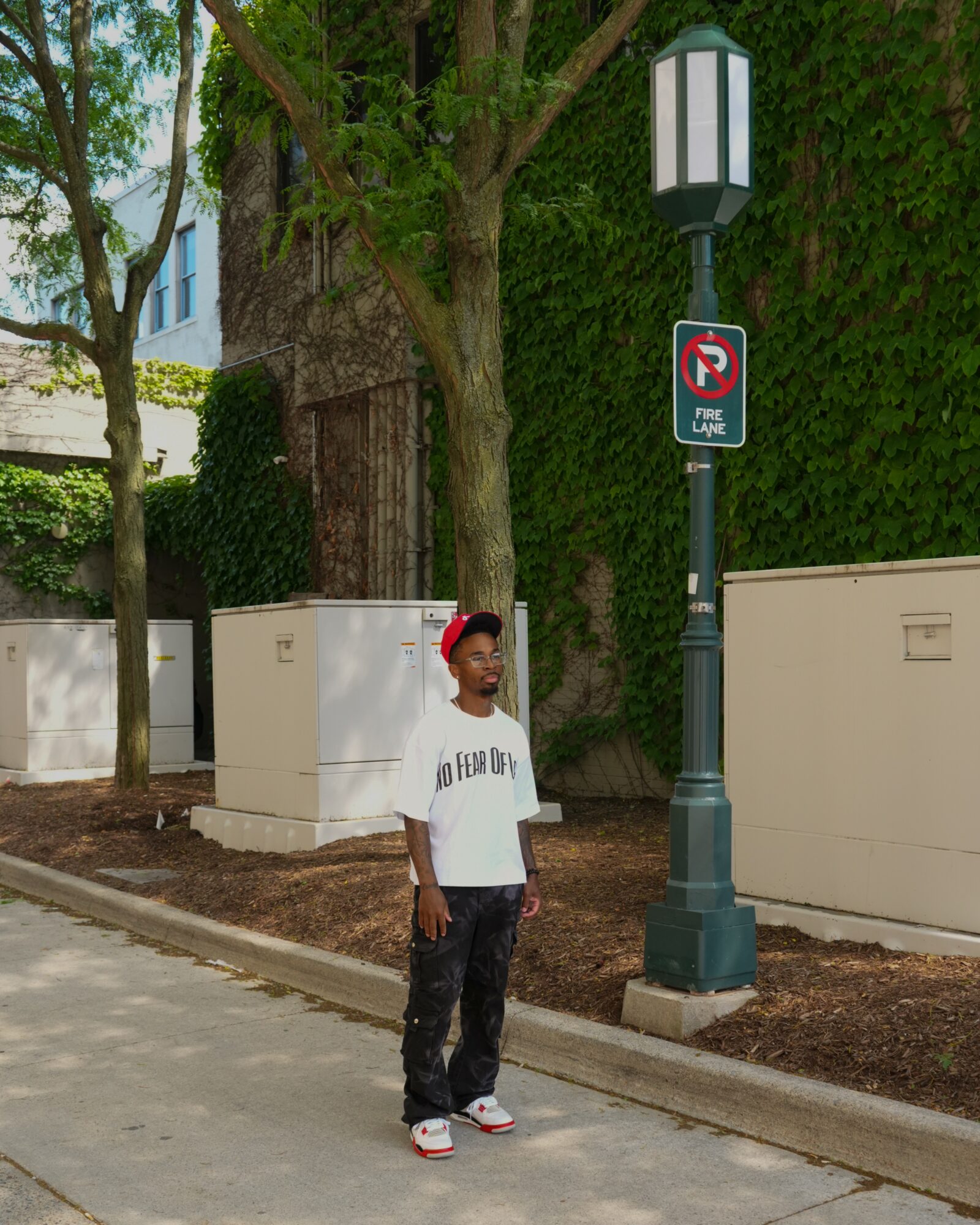 Young person standing on sidewalk near a street lamp and no parking sign, with trees and buildings in background.