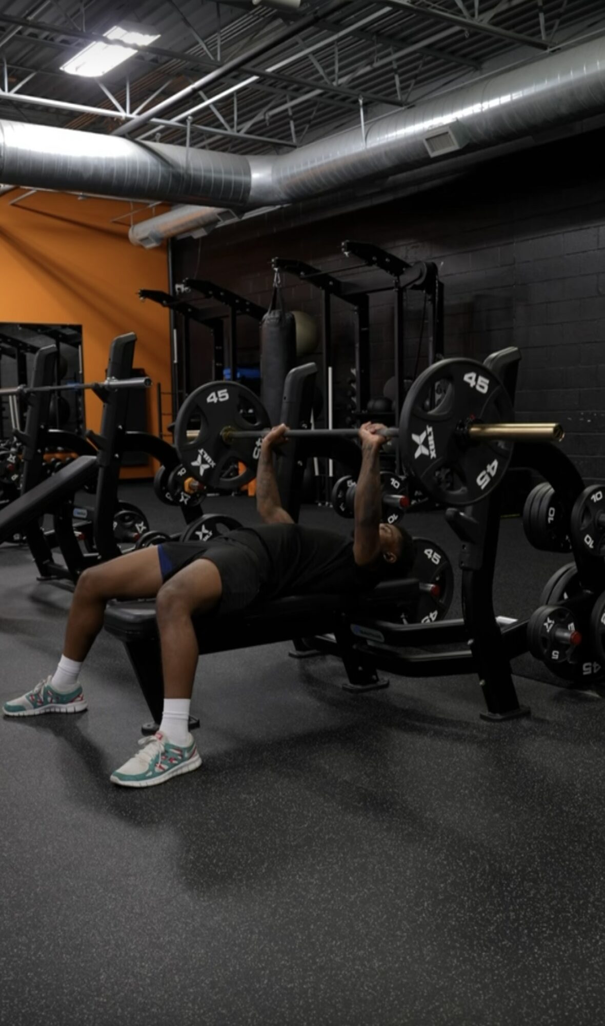 Person lying on a bench press in a gym, lifting weights with a barbell, surrounded by gym equipment and black walls.