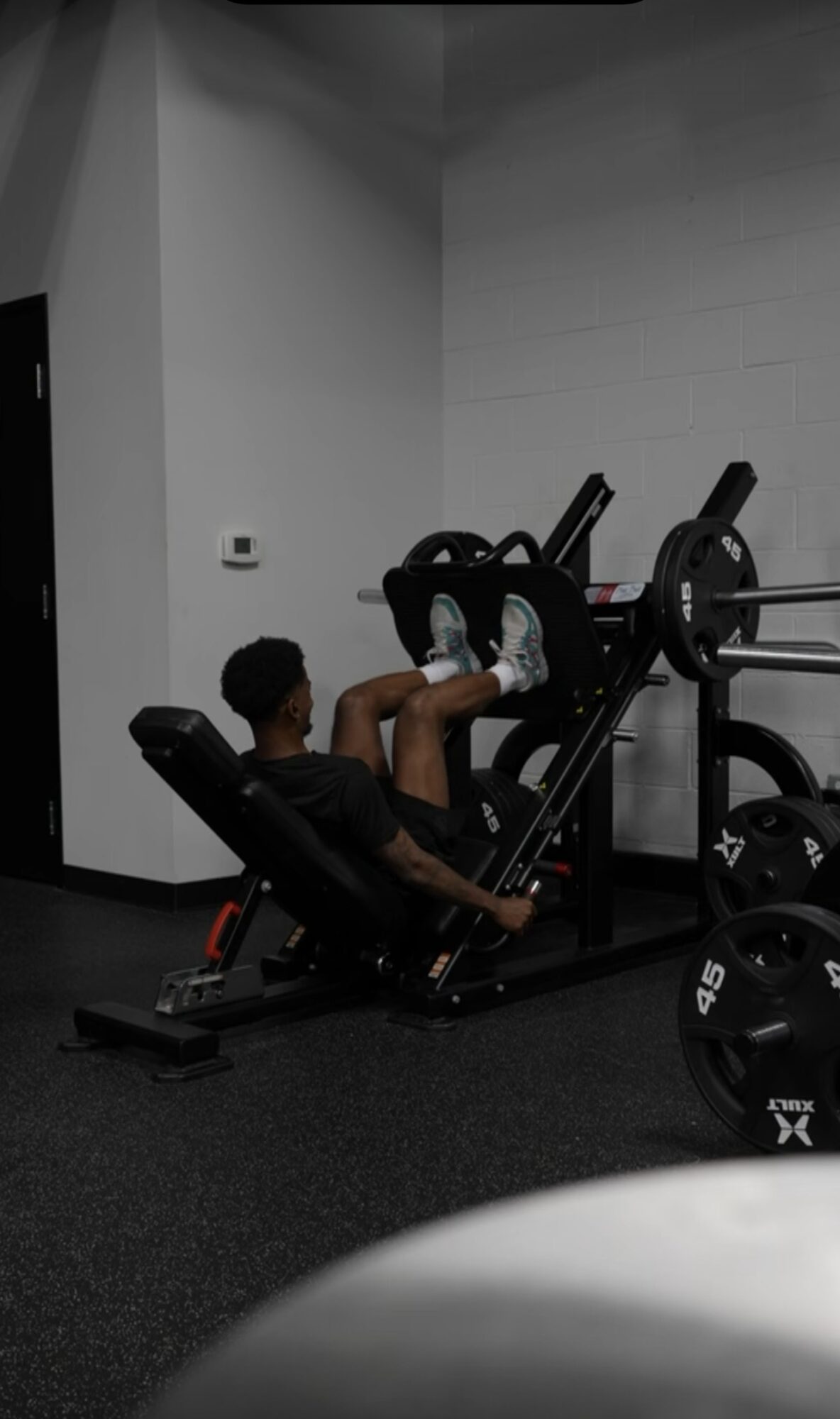 Person sitting on gym equipment, adjusting weights, in a fitness room with white walls and black flooring.