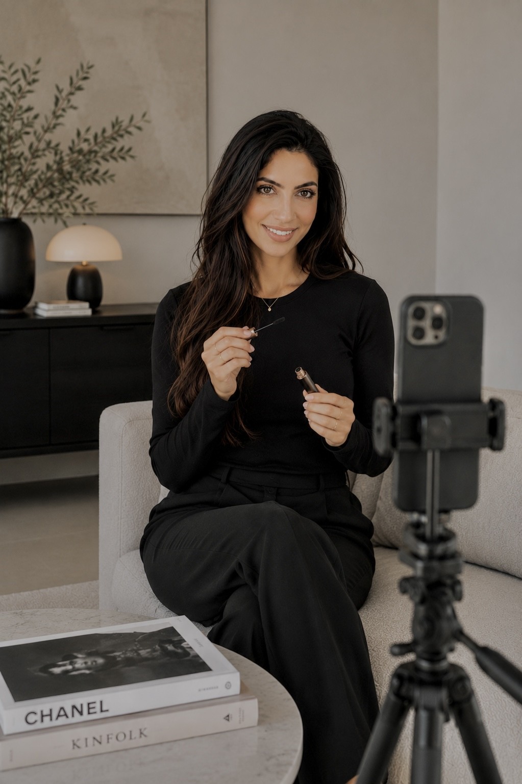 Woman sitting on a chair, smiling, during a video recording, with a camera on a tripod in front, in a modern room.