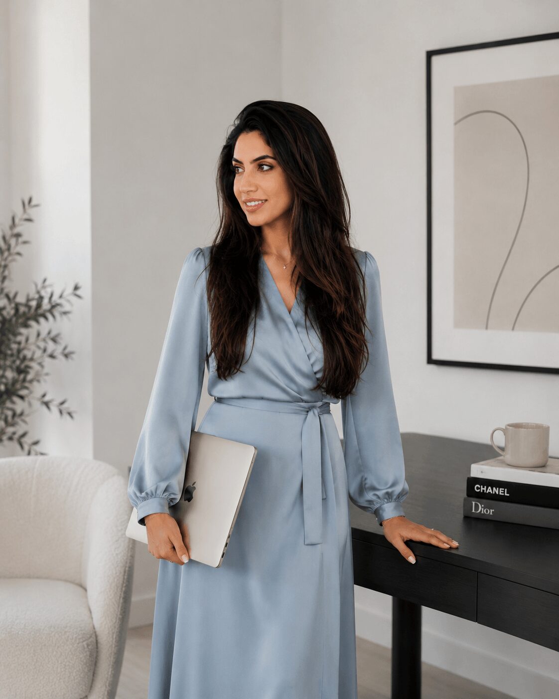 Woman in a light blue dress holding a laptop, standing near a black table with books and a cup, in a modern room.