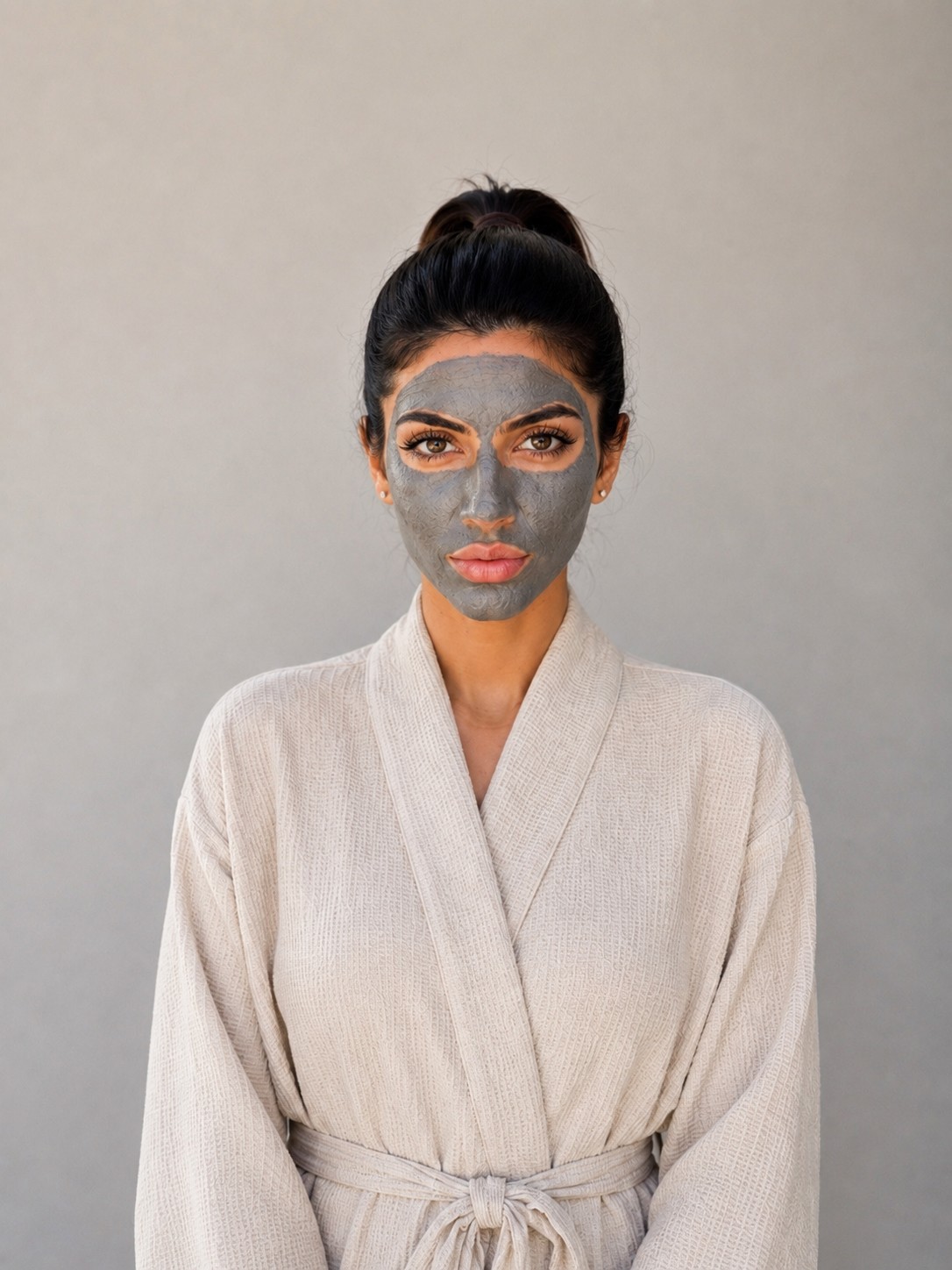 Woman with a facial mask, wearing a light robe, standing against a plain background.