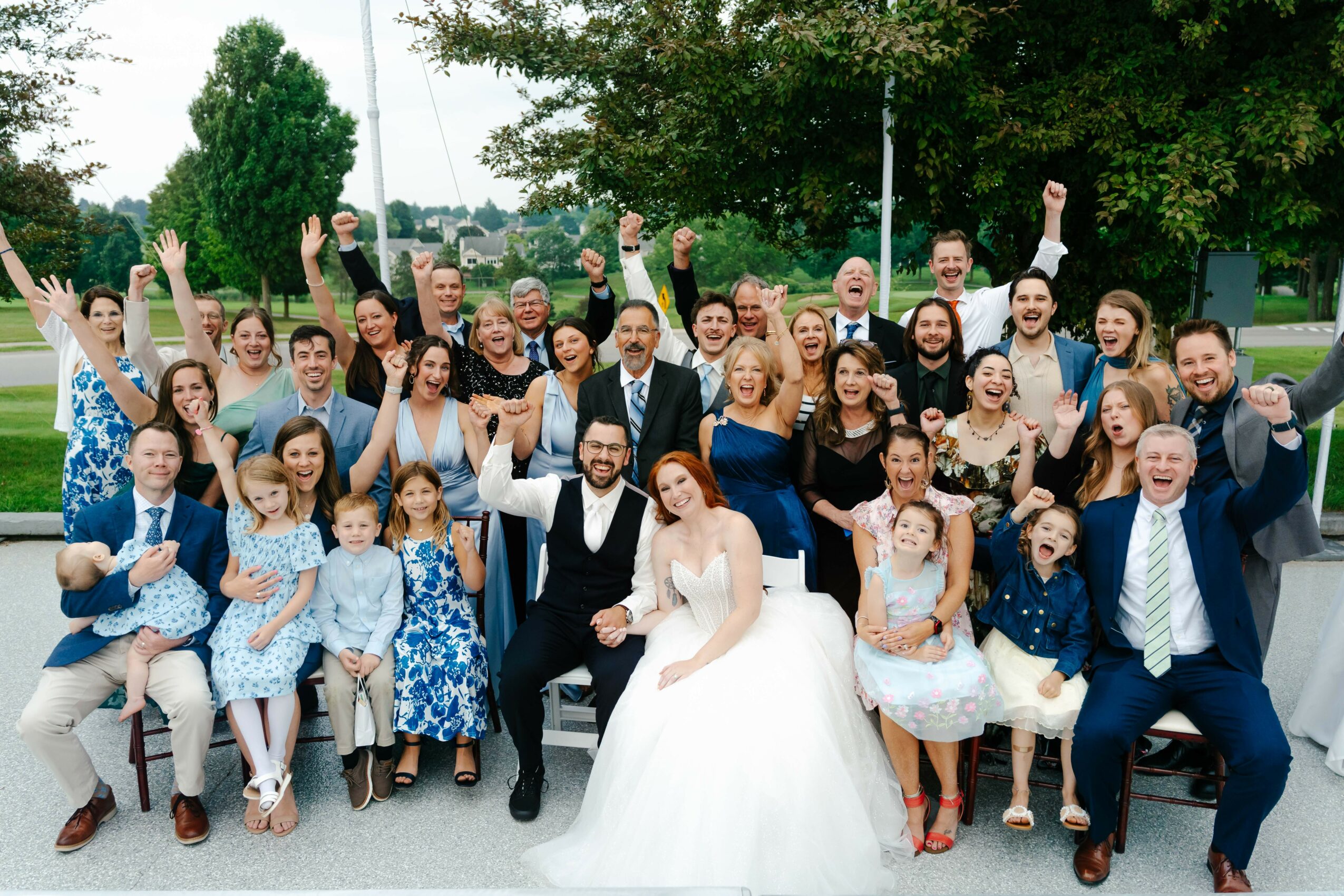 Group of people celebrating outdoors, some raising fists, with a bride and groom in the center, children in front, trees in background.