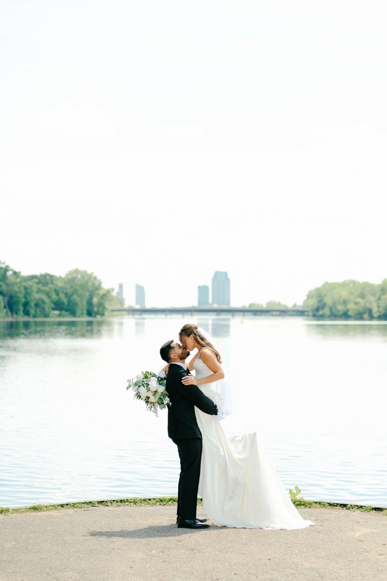 A couple in wedding attire stands by a river, with city buildings in the background, holding each other closely.