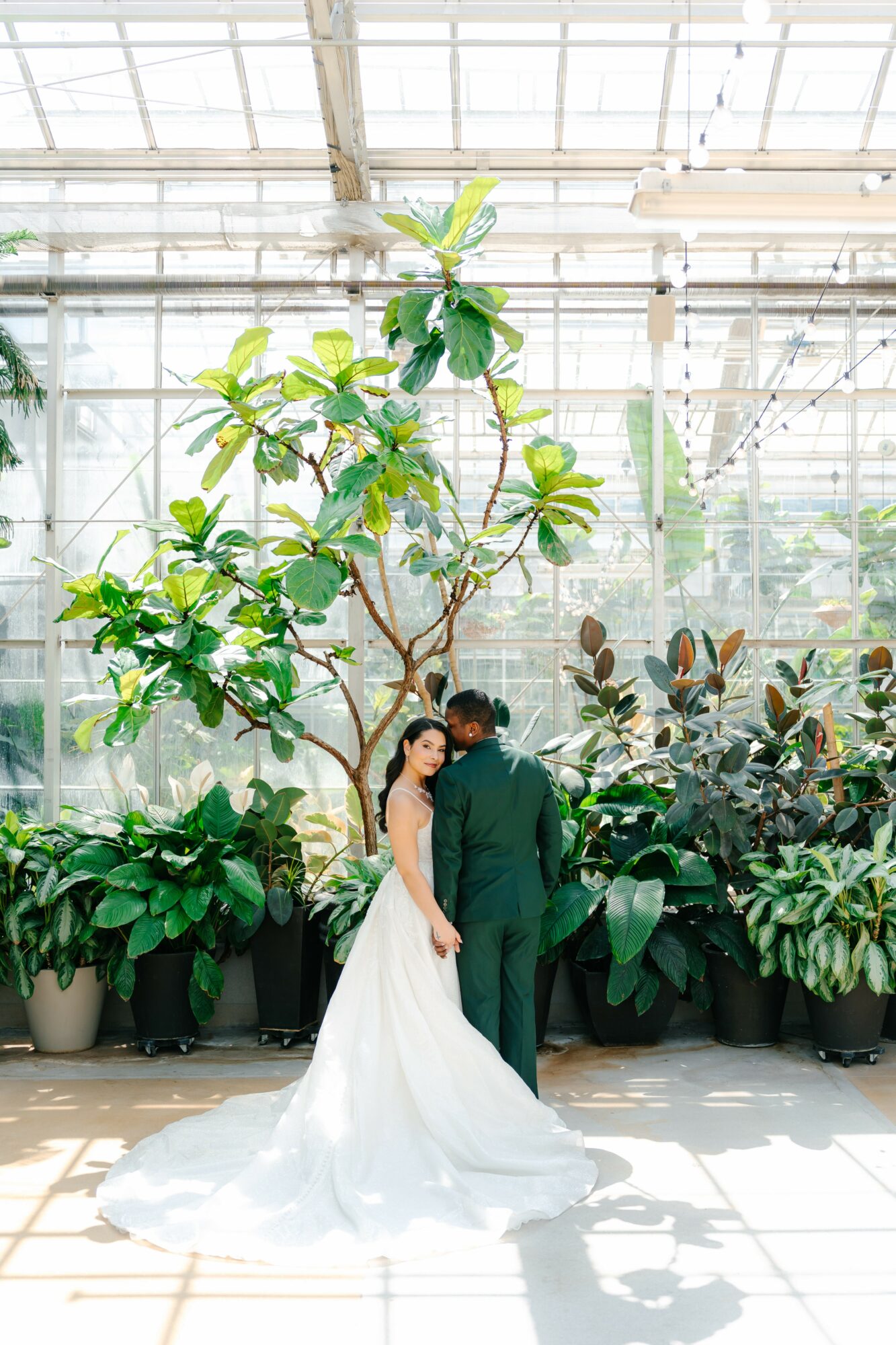 A couple in wedding attire stands close together in a greenhouse with large potted plants and a tall tree behind them.