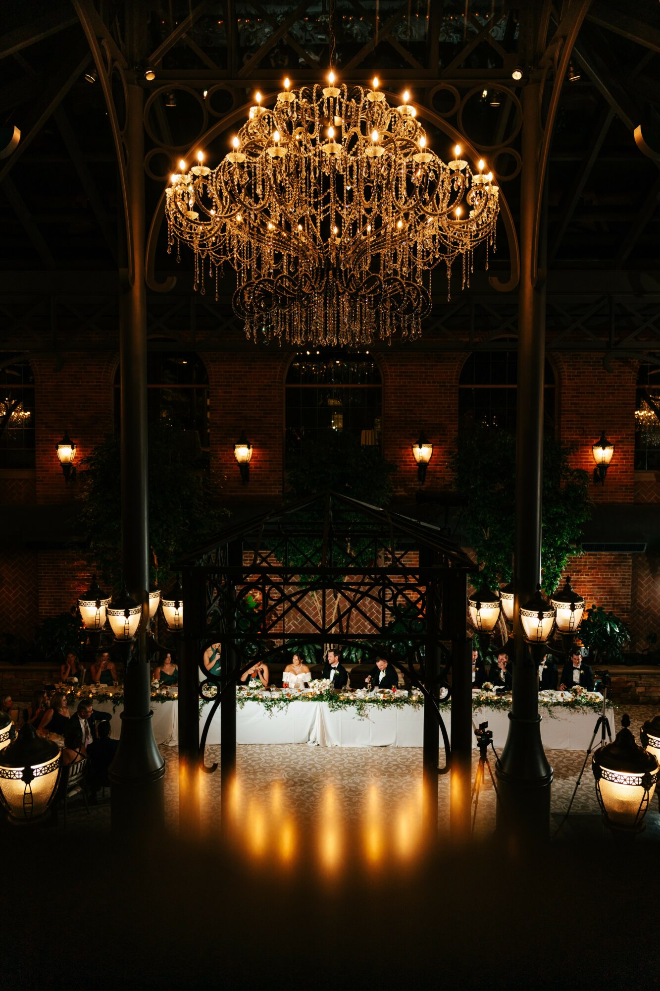 Elegant event space with a large chandelier hanging from a high ceiling, decorated tables, and warm lighting.