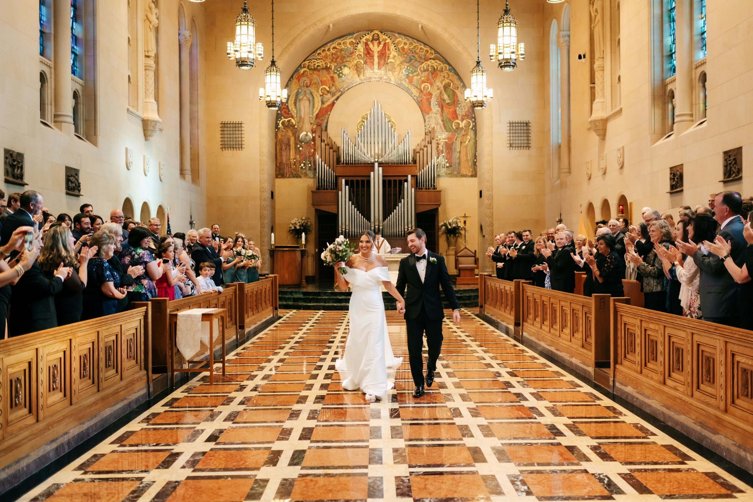 Bride and groom walking down the aisle in a church with guests clapping, ornate ceiling, and large organ in the background.