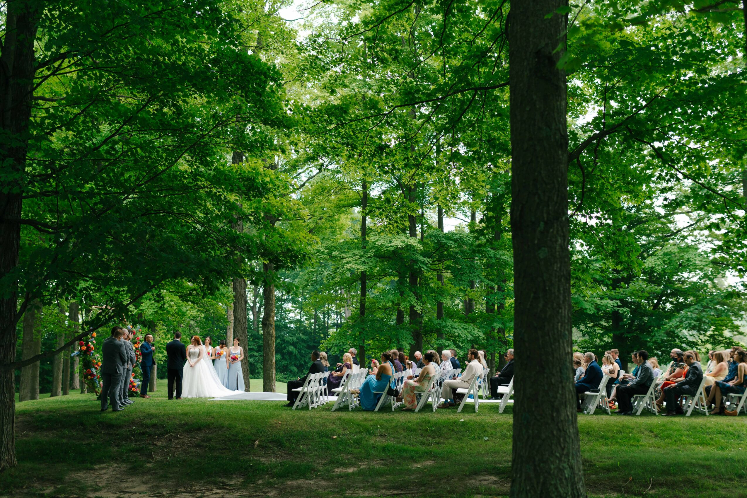 Outdoor wedding ceremony with guests seated in a forested area, bride and groom at altar, trees and greenery around.