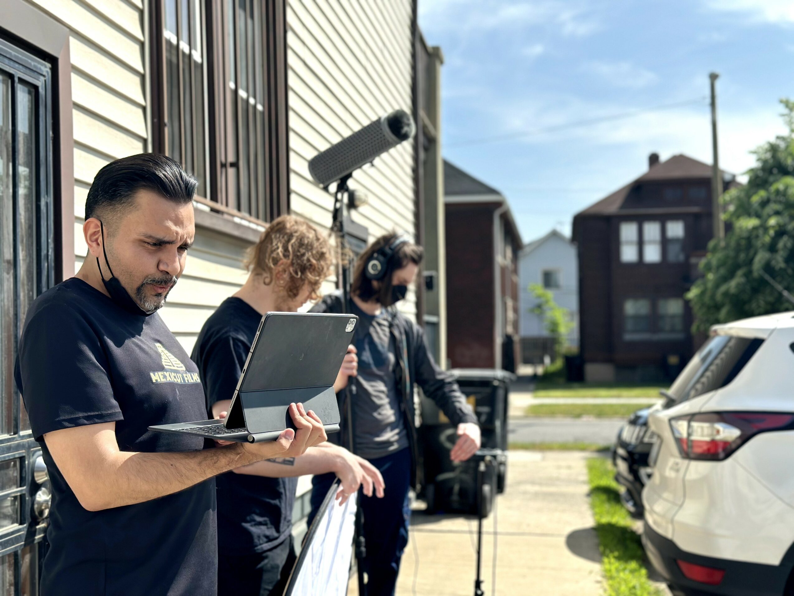 Three people standing outdoors near parked cars, one using a laptop, with buildings and a blue sky in the background.