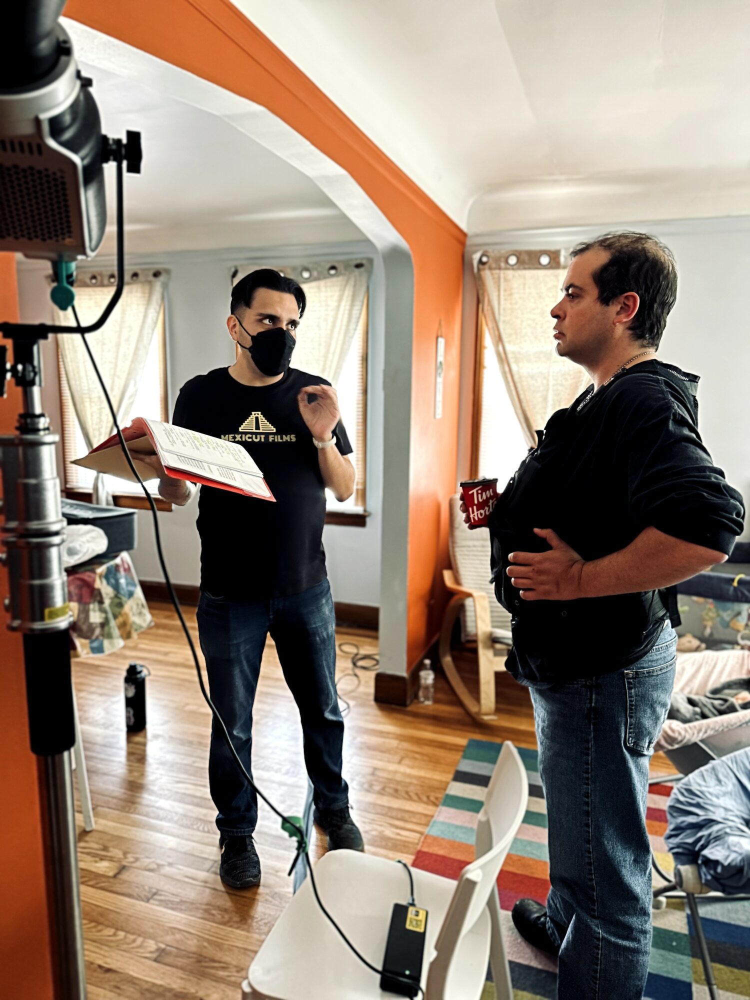 Two men in a living room, one with a mask holding a notebook, the other holding a mug, engaged in conversation.