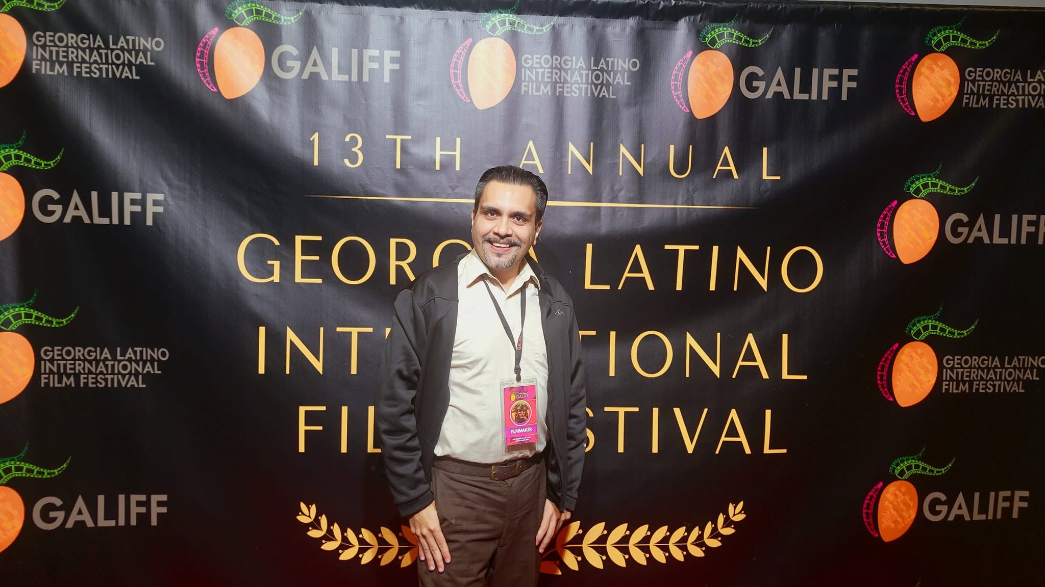 Man standing in front of a black backdrop with festival branding, smiling, wearing a suit and lanyard.