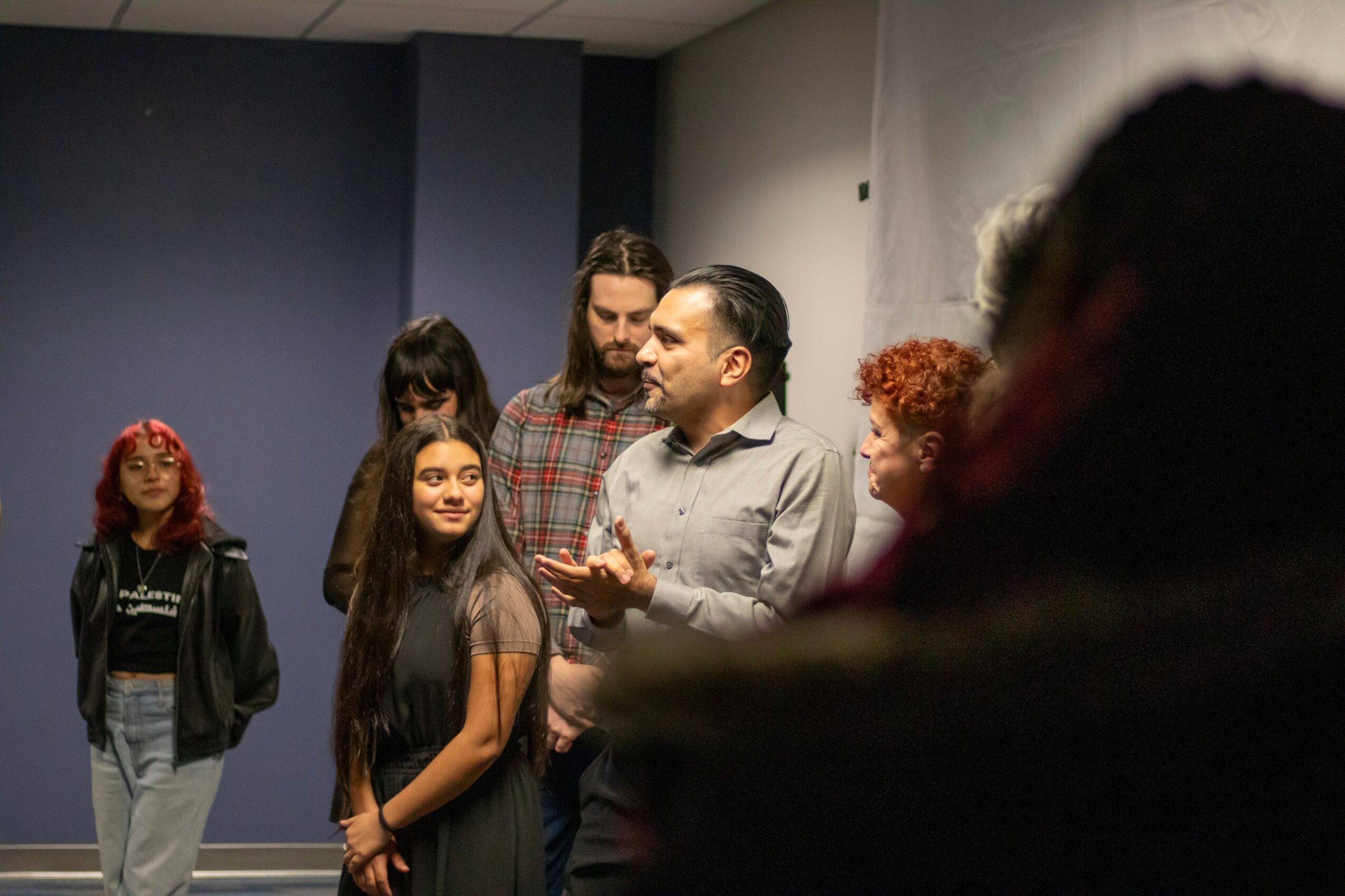 Group of people, including children and adults, gathered indoors, engaged in conversation, with a plain wall background.