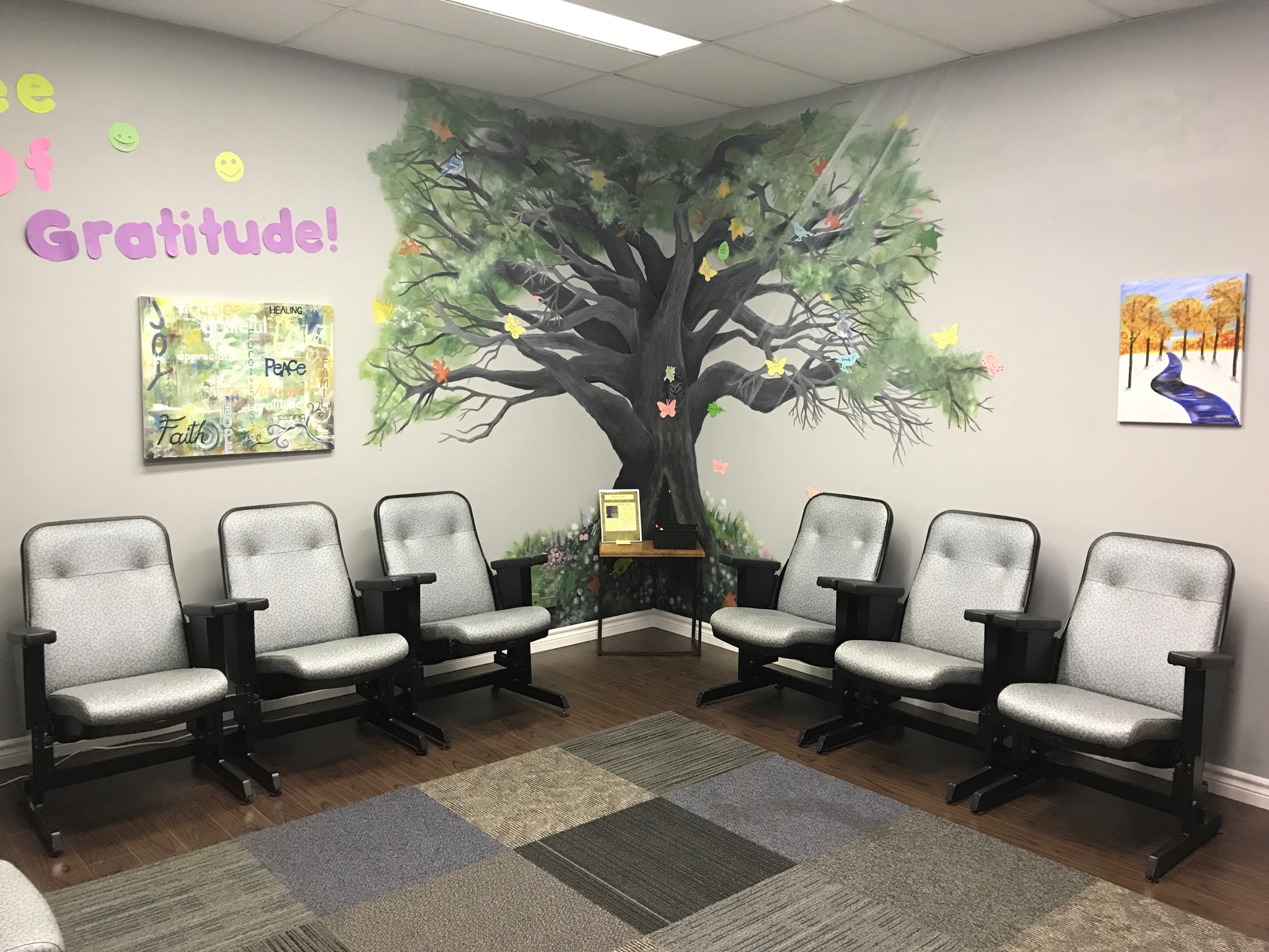 Waiting room with six chairs, a large tree mural on the wall, and framed artwork, with a rug on the floor.