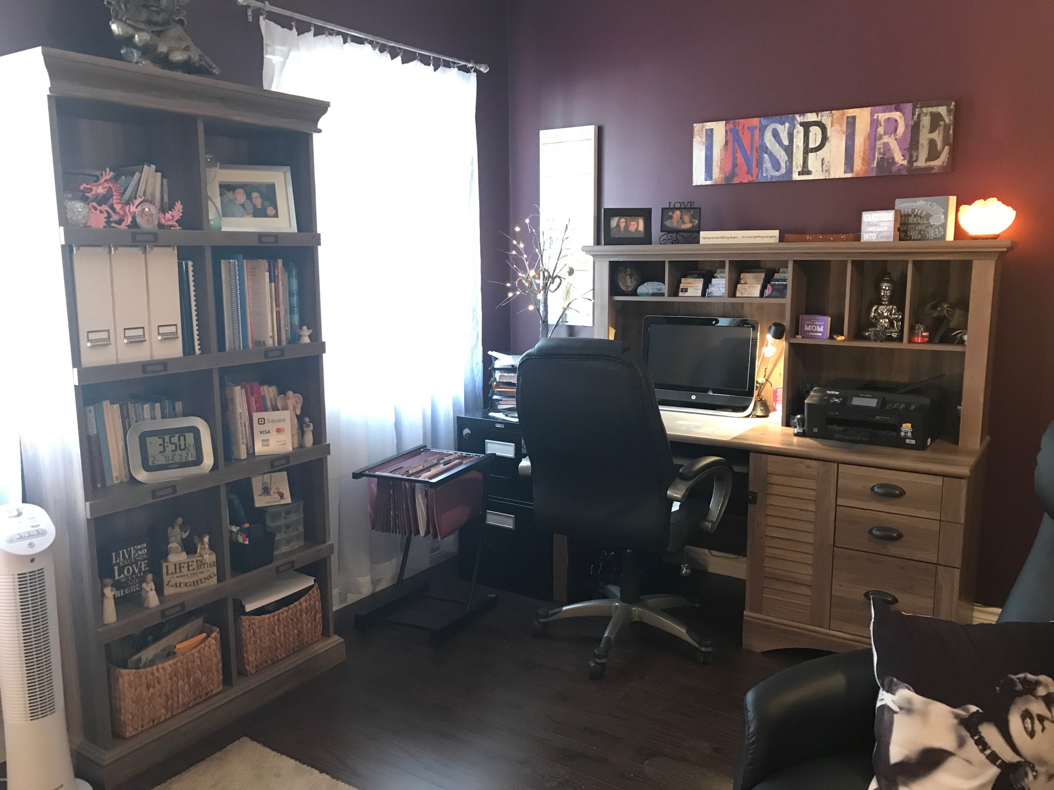Home office with desk, bookshelf, and window with curtains, dark purple wall, and a black chair.
