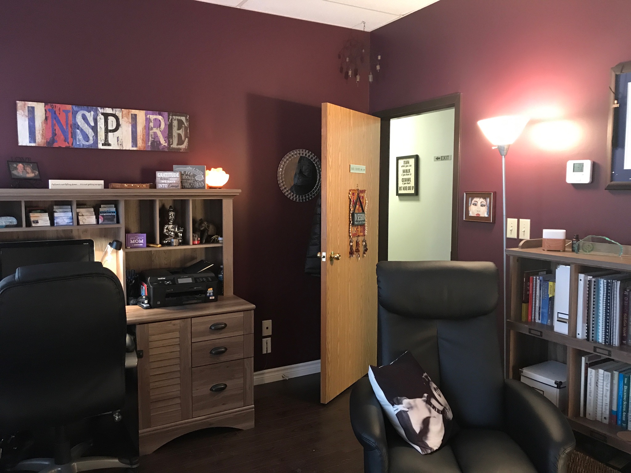 Office room with maroon walls, bookshelf, desk, and a black chair. Open door reveals a hallway. Wall decorations include a clock and framed pictures.