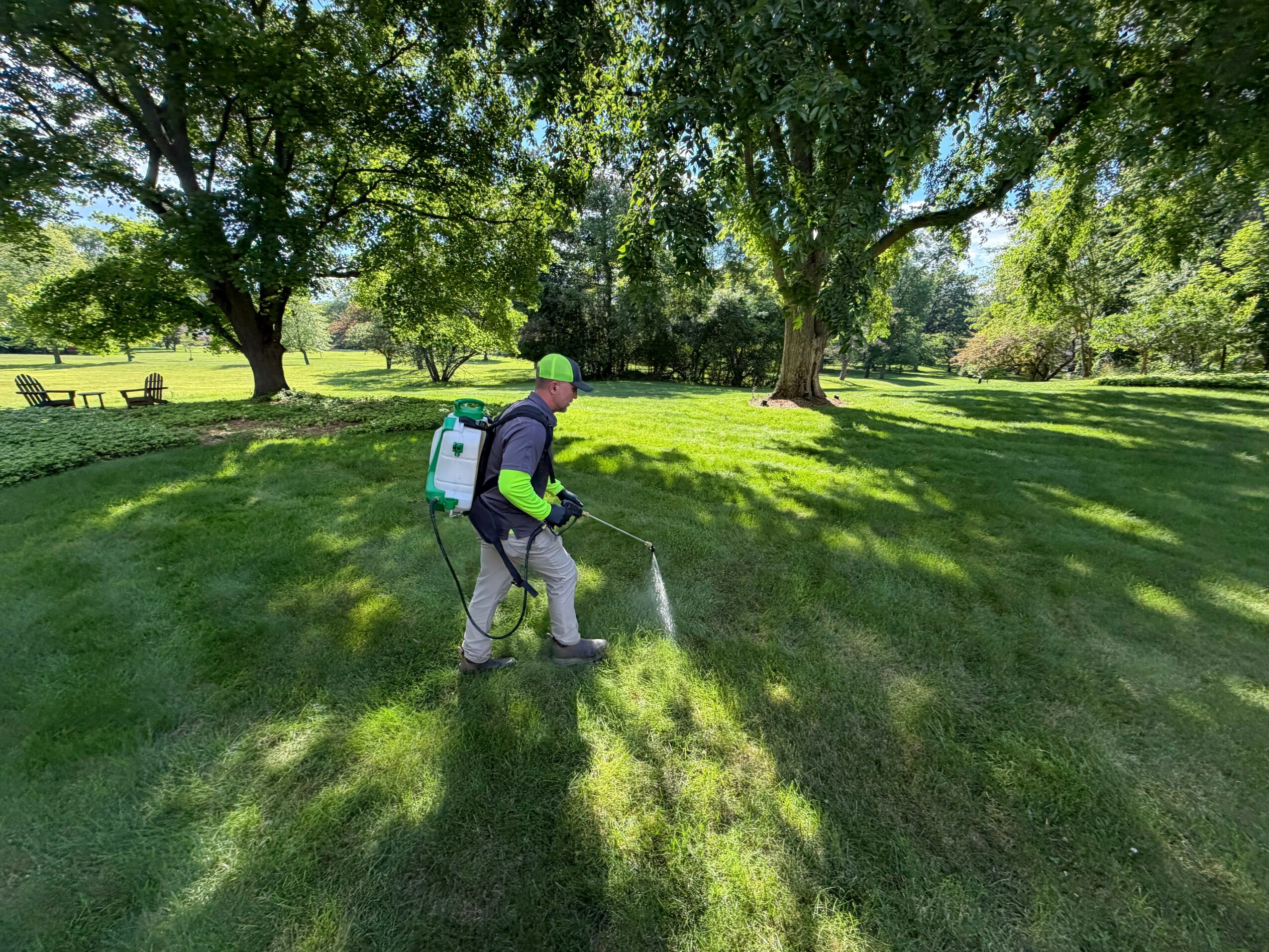 Person wearing a backpack and spraying water on grass in a park with trees and benches, sunny day.