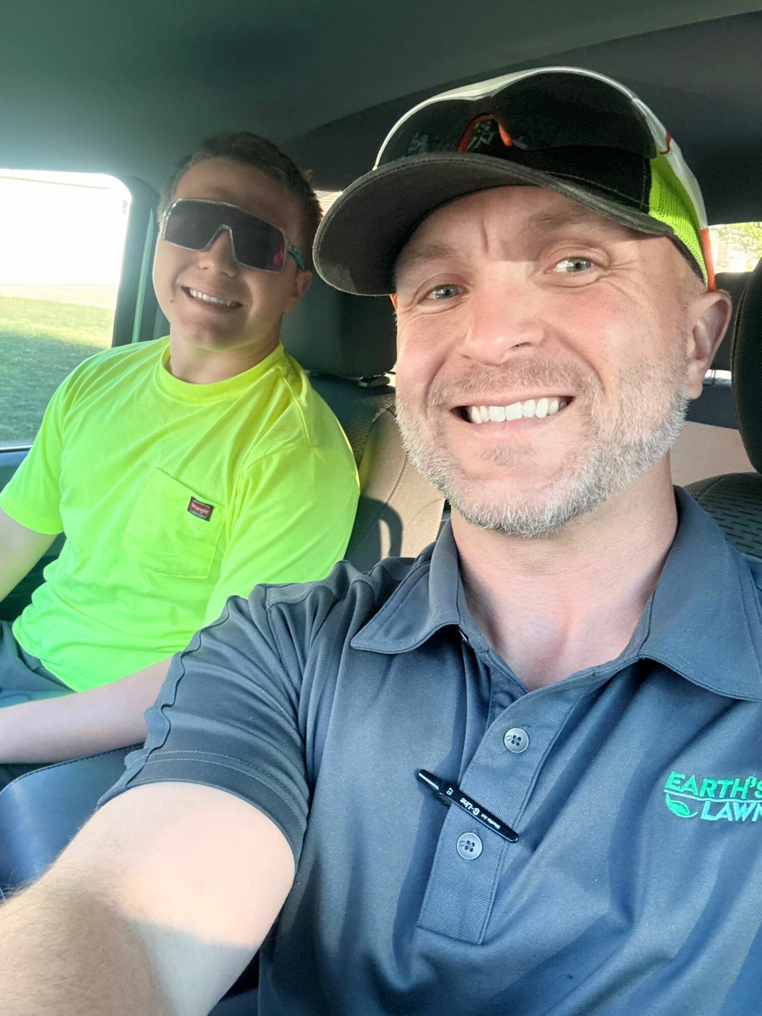 Two men sitting in a vehicle, smiling at the camera; one wearing a blue shirt and cap, the other in a bright yellow shirt.