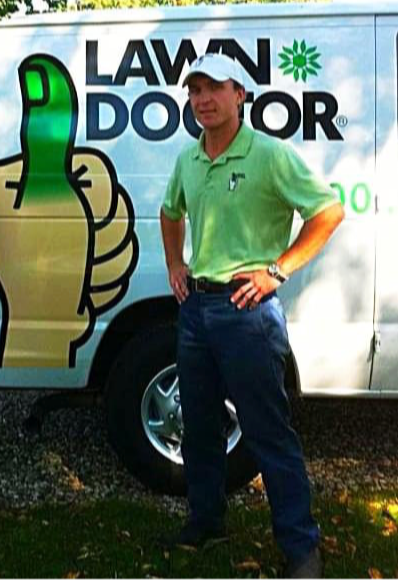 Man in green polo shirt and cap standing in front of a lawn care vehicle with a thumbs-up sign, outdoors.