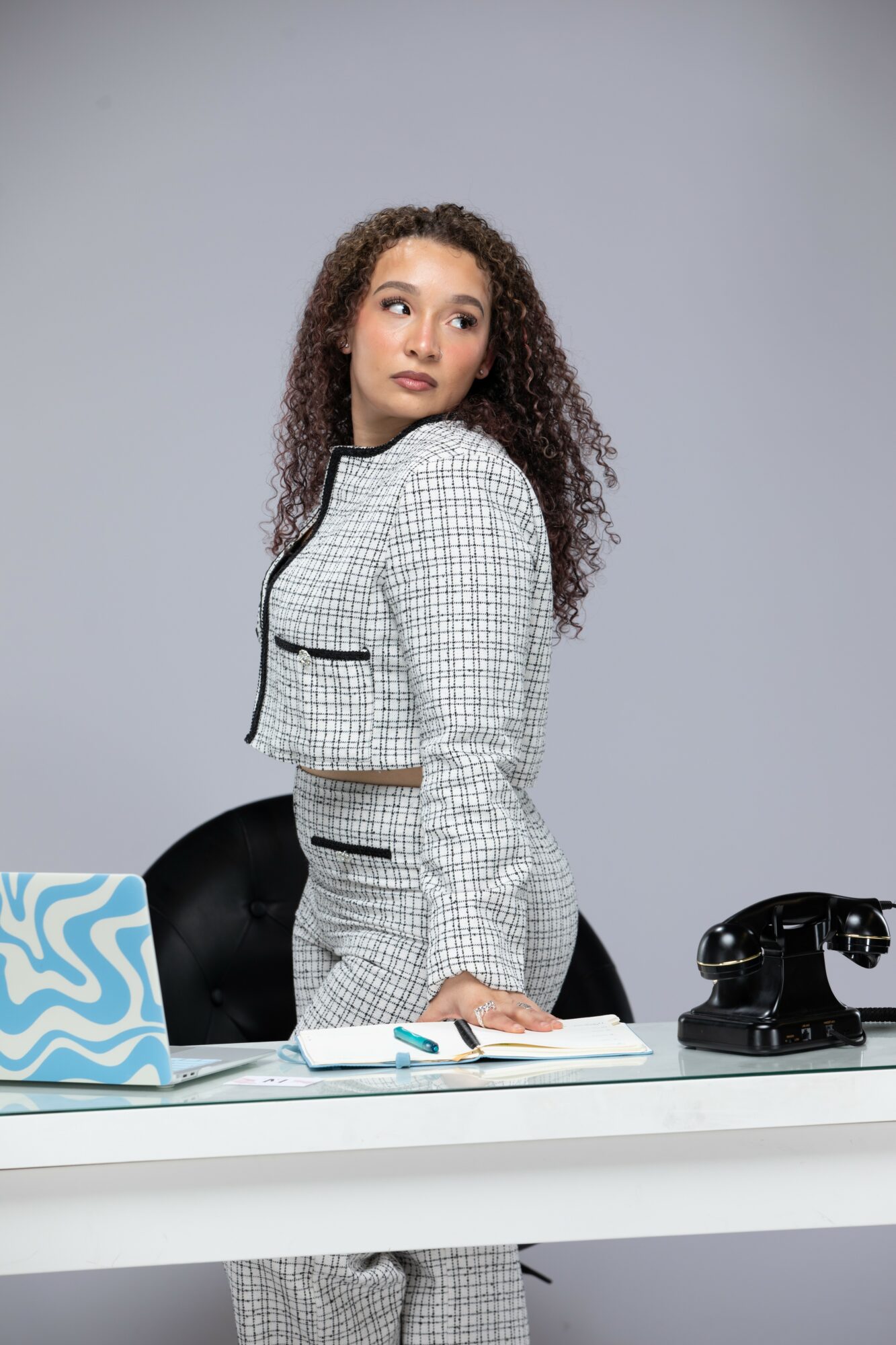 Young woman with curly hair in a checkered blazer standing behind a desk with a laptop and a rotary phone, looking to the side.