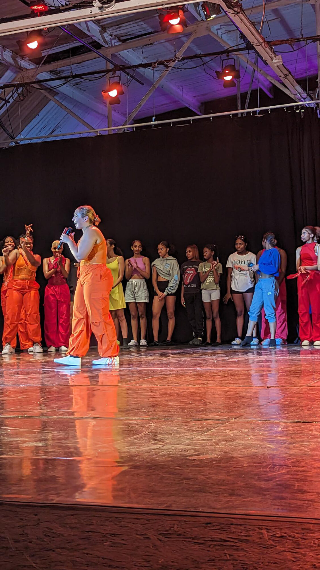 Group of performers and children on stage with black curtain background, colorful costumes, and stage lighting.