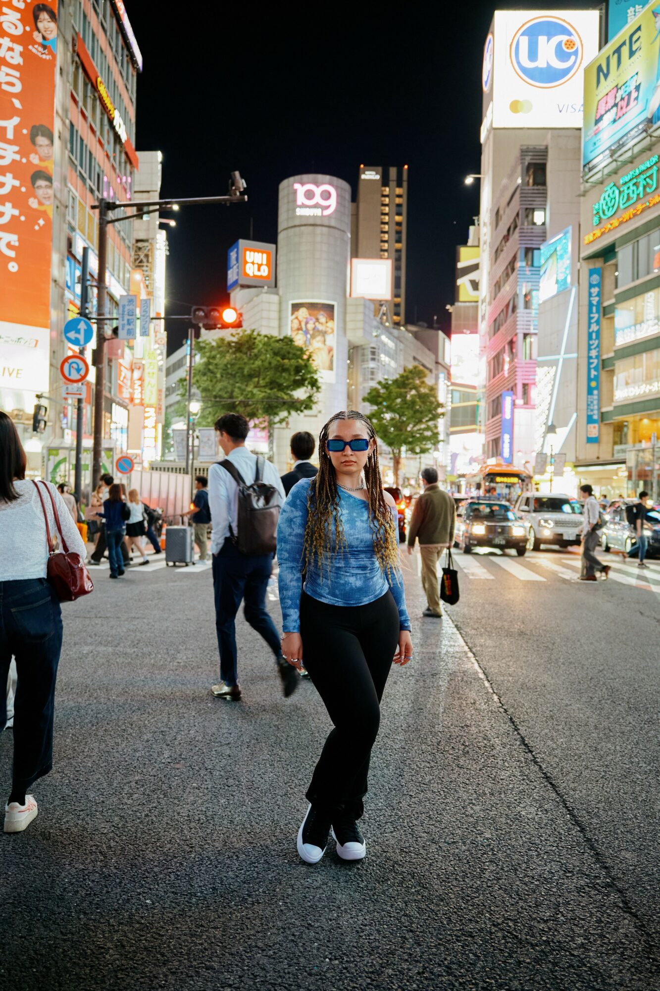 Woman walking on busy city street at night, illuminated billboards and buildings in background, wearing sunglasses and blue top.